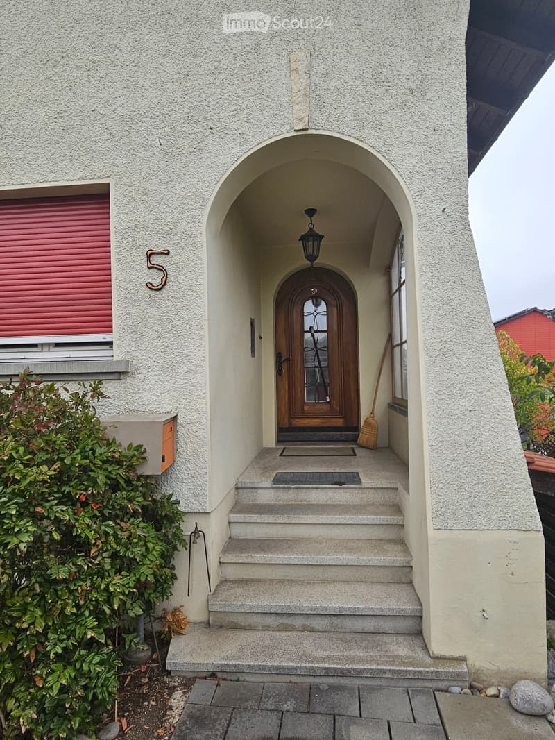 Front entrance of a house with a stone facade, arched entryway, wooden double doors, and a small set of steps leading up to the door.