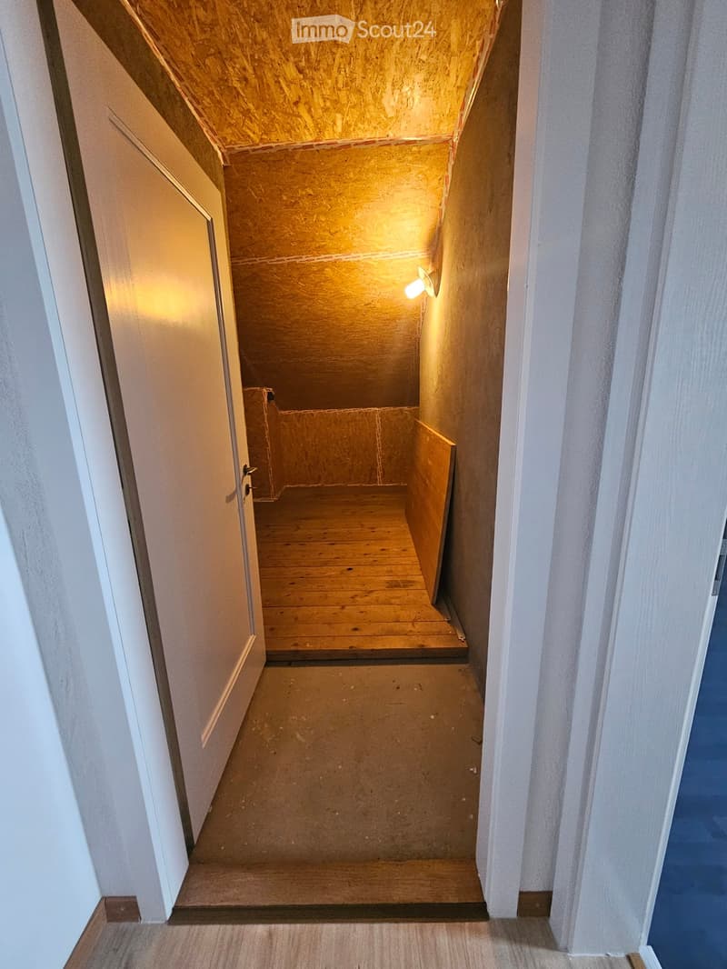 Narrow, unfinished hallway with wooden stairs leading to an attic space. Exposed wooden beams and walls, warm lighting from a wall-mounted lamp.
