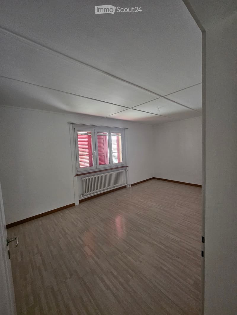 Empty room with wooden flooring, white walls, and a window with blinds. A radiator is placed below the window.