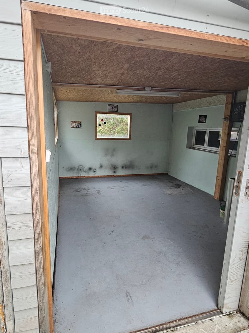 Empty room with concrete floor, light blue walls, and wooden ceiling beams. Features a small window and a partially open door leading to another area.