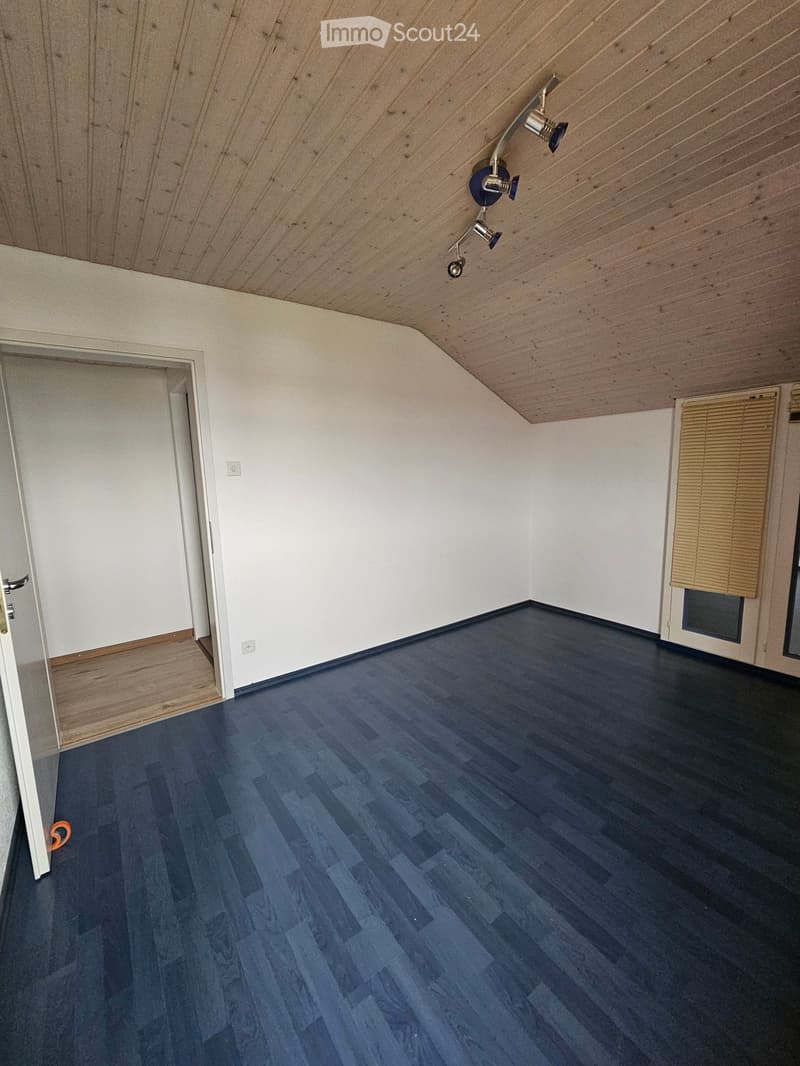 Empty room with dark blue wooden flooring, white walls, and a slanted wooden ceiling. Features include a door on the left and a window with blinds on the right.