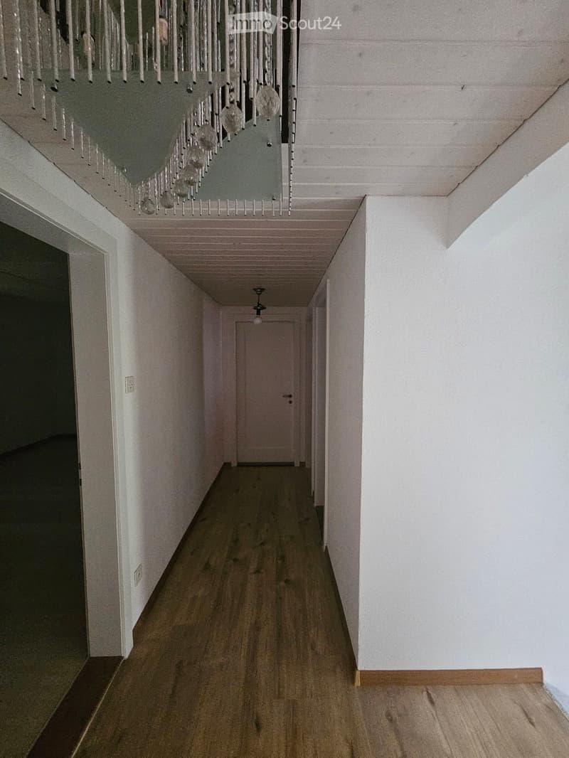A long, narrow hallway with wooden flooring, white walls, and a chandelier hanging from the ceiling. The hallway leads to a closed door at the end.