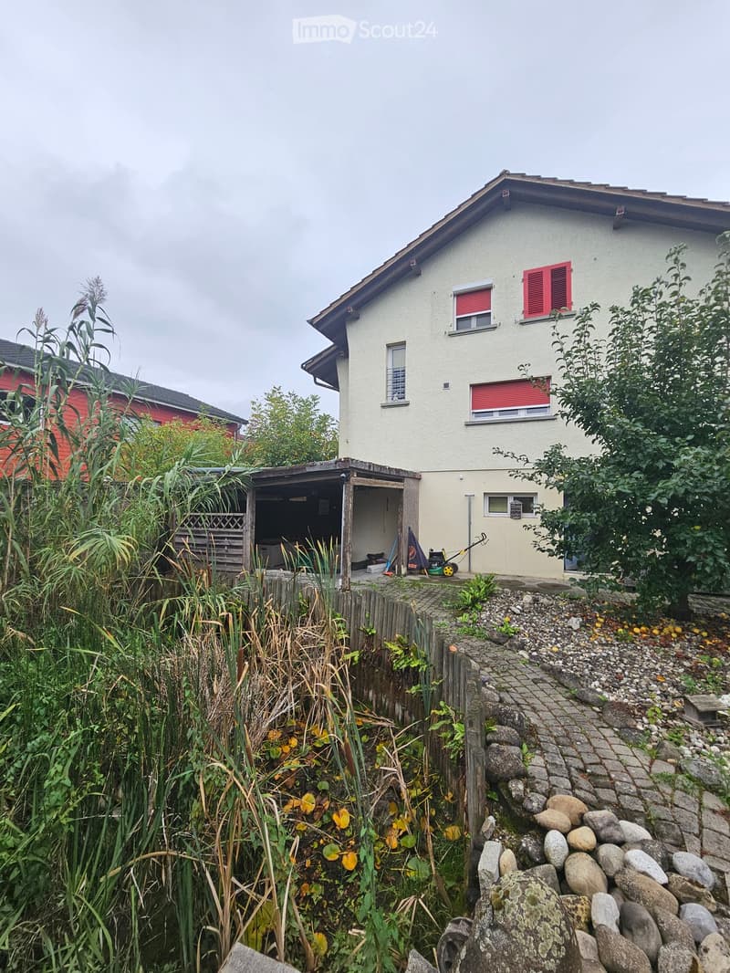 Two-story house with beige exterior, red shutters, and a small covered outdoor area with tools and a tarp.