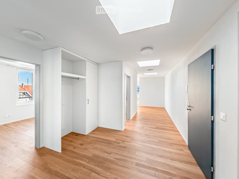 Bright, minimalist hallway with wooden flooring, white walls, and recessed lighting. Features built-in shelving unit and a window offering a view of the exterior.
