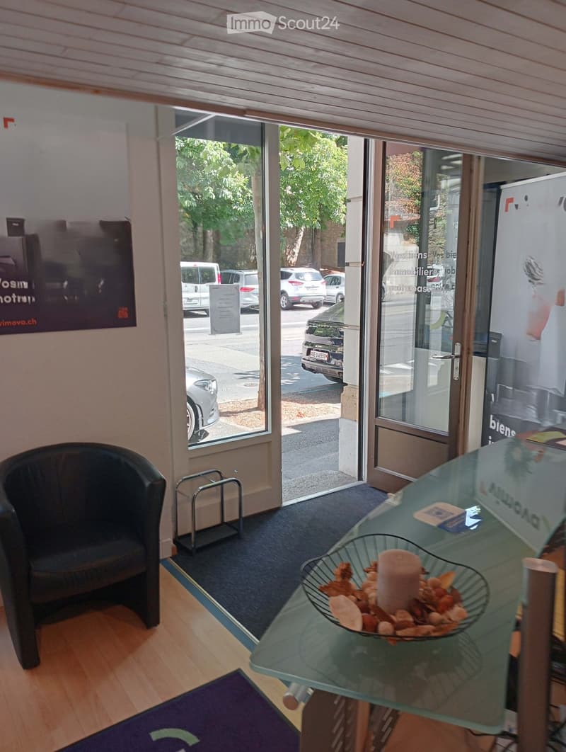 A small, well-lit office or reception area with a wooden floor, a glass table with a decorative bowl, a single chair, and a small metal stand near the entrance.