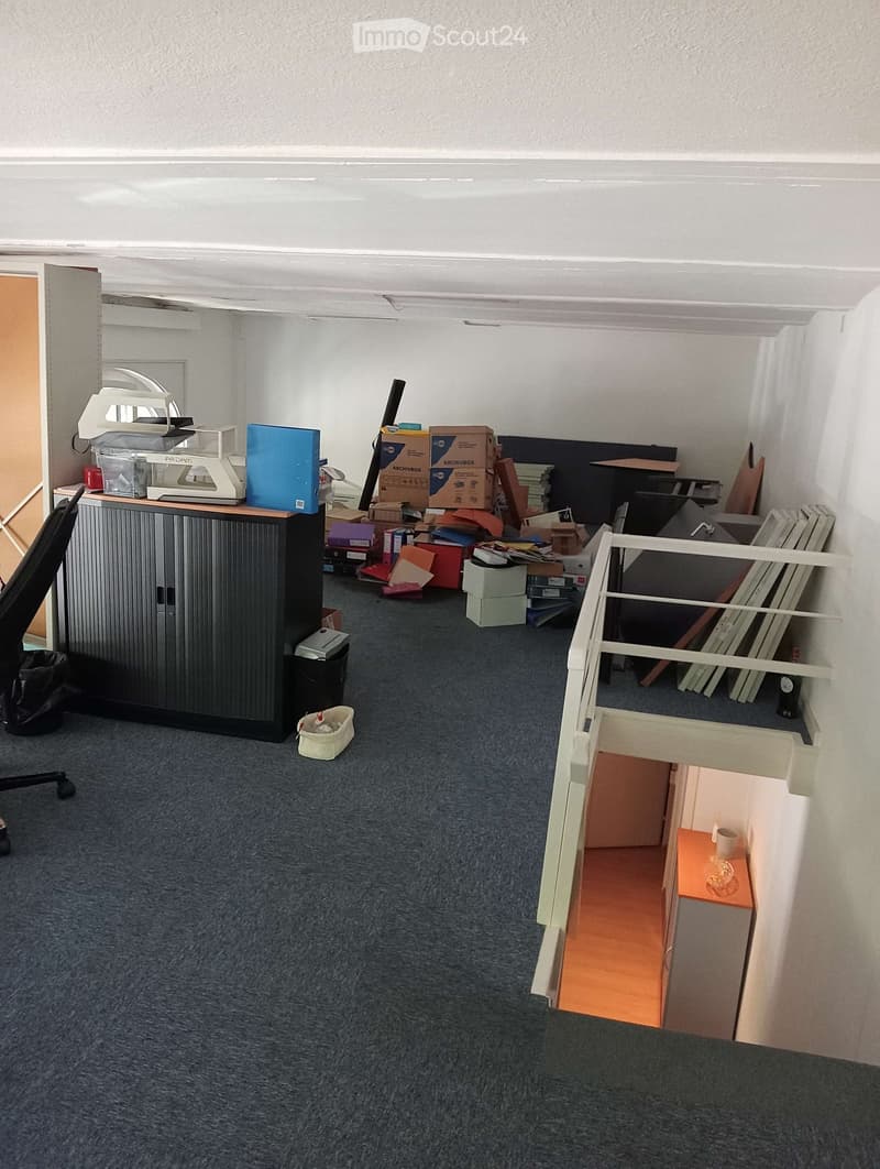 The room has a grey carpeted floor, white walls, and a black cabinet with a printer on top. There is a small white stool with a cup on it, a black chair, and several boxes and items scattered around. A small staircase leads to another area with a small ta