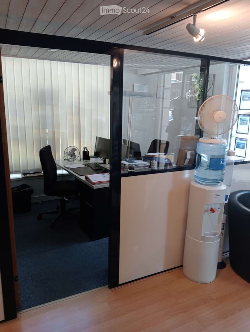 Office space with a desk, office chair, computer monitor, keyboard, water cooler, and fan. The room has glass partitions and a wooden floor.