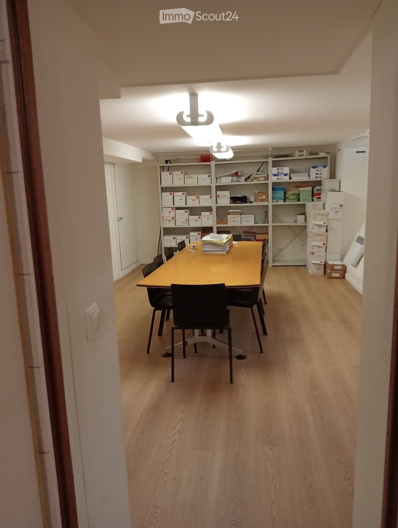 A small, well-lit room with wooden flooring and a large table surrounded by chairs. Shelves with boxes and files are visible in the background.