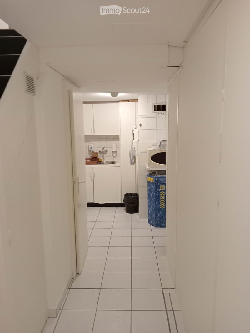 A narrow, white-tiled hallway leading to a small kitchenette area with white cabinets, a sink, microwave, and a trash bin.