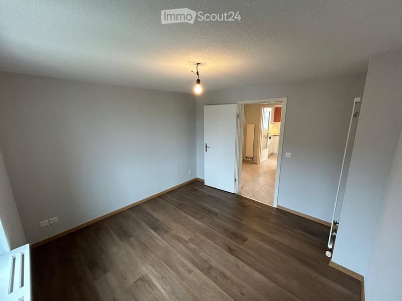 Empty room with wooden flooring, light grey walls, and a single hanging light fixture. An open doorway leads to another room with tiled flooring and white cabinets.