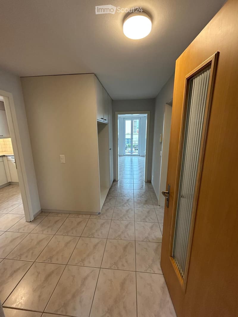 Empty hallway with tiled flooring, white walls, and a round ceiling light. A wooden door is visible on the right, and an open doorway at the end leads to another room.