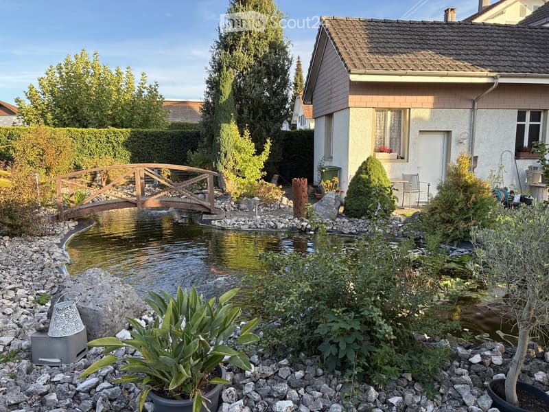 Garden with a small pond, wooden bridge, surrounding greenery, and a house in the background.