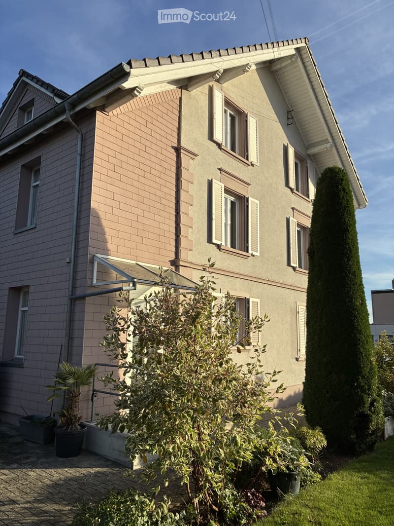 Two-story house with beige siding, multiple windows with shutters, and a well-maintained garden with potted plants and shrubs.