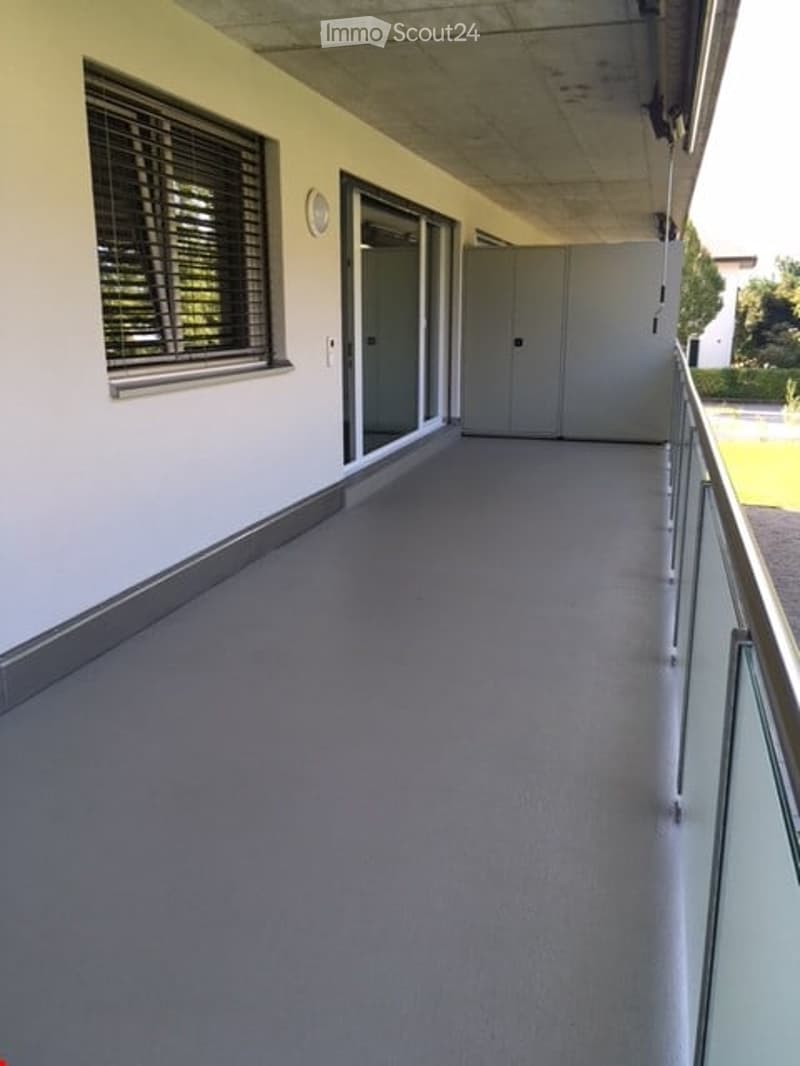 Empty balcony with grey flooring, glass railing, sliding doors, and a window with blinds.