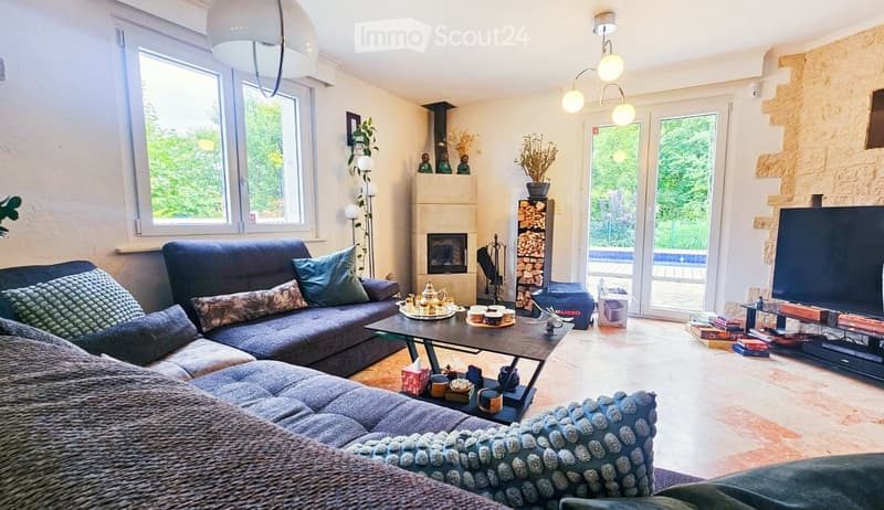 Bright living room with large windows, a dark blue sectional sofa, a glass coffee table, a fireplace, and a television. The room features a modern chandelier and a view of a garden through large glass doors.