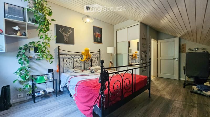 Cozy bedroom with a metal-framed bed, red and white bedding, nightstand with lamp, potted plant, mirror, and shelves with decorative items.