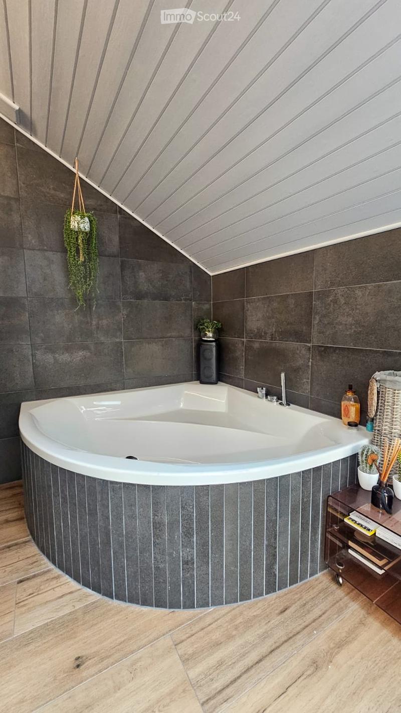 Modern bathroom with a large, curved bathtub, dark grey tiles, wooden flooring, and decorative elements including a hanging plant, a speaker, and a small shelf with books and candles.