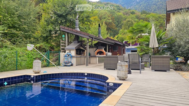 Outdoor area with a swimming pool, wooden decking, outdoor seating, and a barbecue area surrounded by lush greenery and trees.