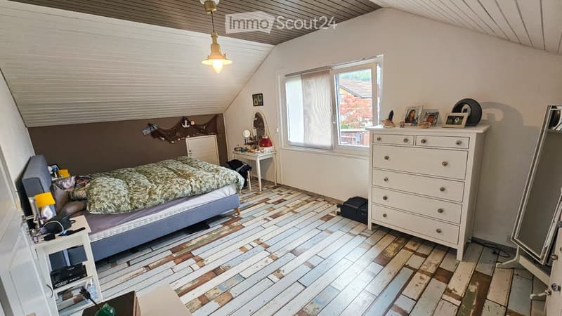 Cozy attic bedroom with slanted ceilings, wooden flooring, a bed with a patterned duvet, a nightstand, a dresser, a small table with a mirror, and a window with blinds.