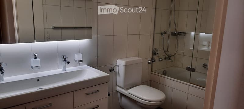 Modern bathroom with a white sink, toilet, and glass-enclosed shower. Features a large mirror with built-in lighting and a sleek faucet.