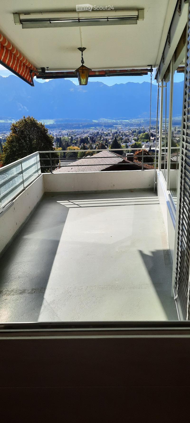 Balcony with a panoramic view of a city and mountains, featuring a glass railing and a hanging light fixture.