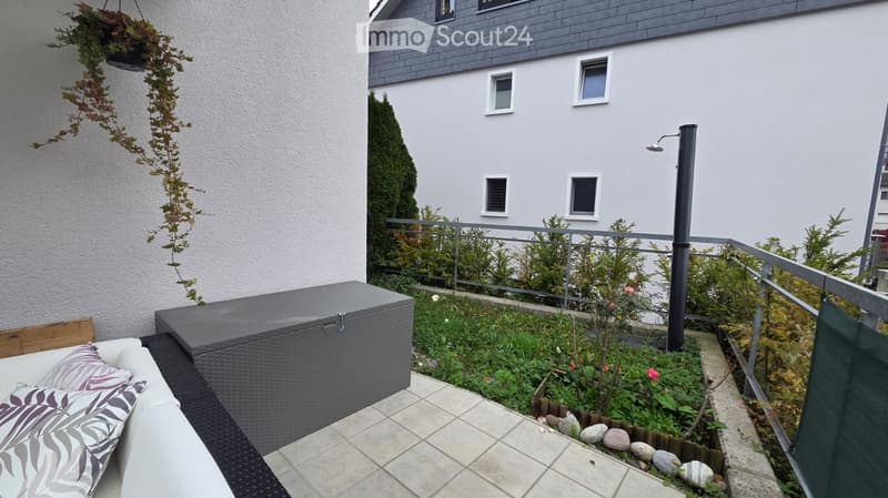 Balcony with a small garden area, featuring a sofa with cushions, a storage box, plants, and a railing with a view of the building.