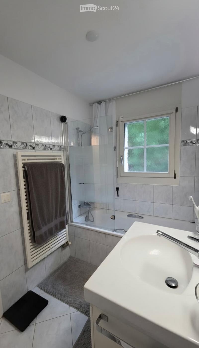Modern bathroom with a white sink, bathtub, shower, and a towel on a radiator. The room features tiled walls and a window with a curtain.