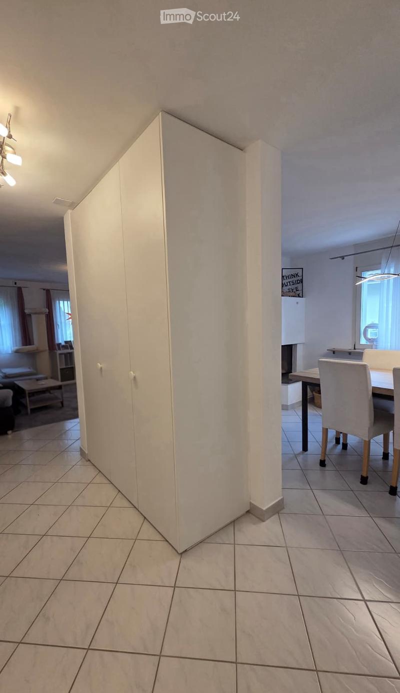 A modern, well-lit hallway with tiled flooring, white walls, and built-in cabinets. Visible furniture includes a dining table with chairs, a small stool, and a lamp.