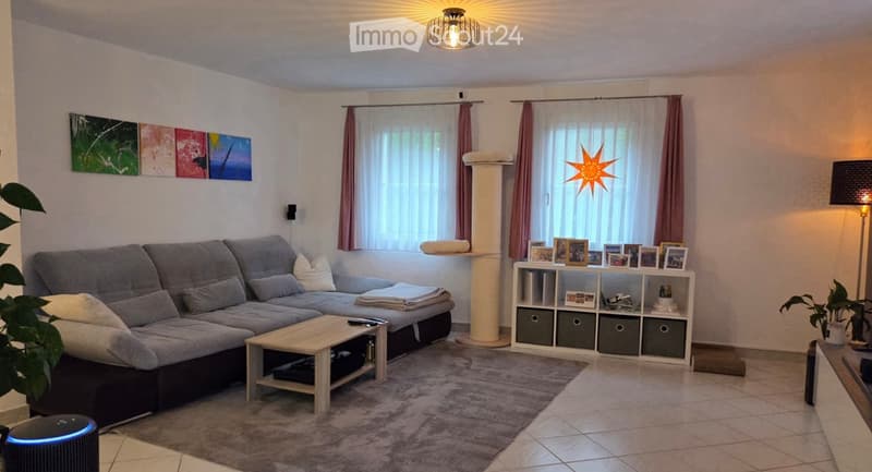 Modern living room with grey sectional sofa, wooden coffee table, white shelving unit with picture frames and storage boxes, floor lamp, table lamp, cat tree, and decorative plants.