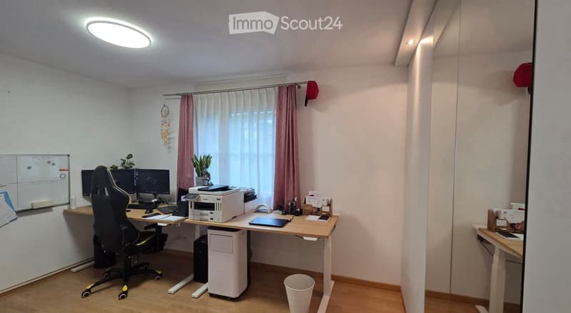 A modern office space with a desk, ergonomic chair, computer, printer, and a large window with curtains. The room features a whiteboard, potted plants, and a trash bin.