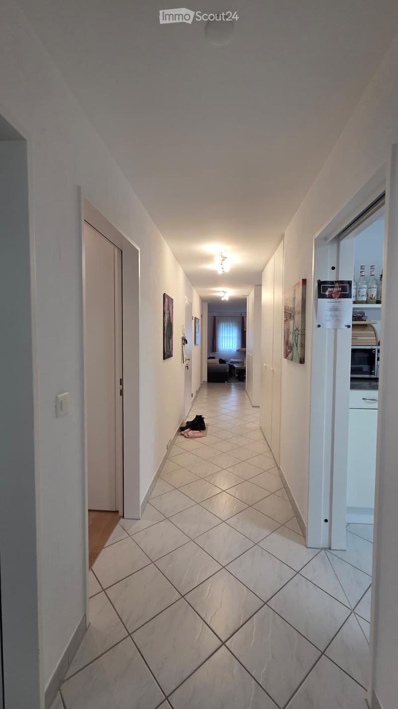 A clean, well-lit hallway with white tiled flooring and white walls. Doors and framed artwork are visible along the walls, and a small area with shoes is placed near one wall.