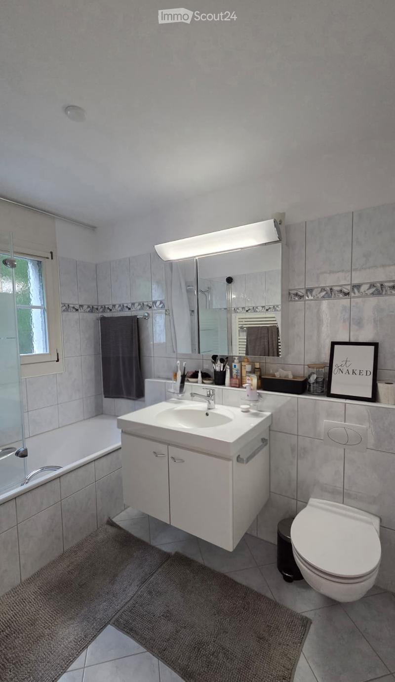 Modern bathroom with a white sink, mirror, toilet, bathtub, and various bathroom accessories. The space features tiled walls and a grey rug on the floor.