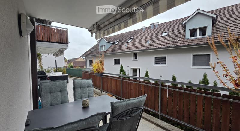 Balcony with a table, chairs, and a vase, overlooking a residential area with houses and greenery.