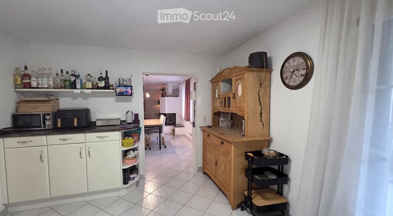 The kitchen features white cabinets, a microwave, a printer, a wooden cabinet with glass doors, a small table with chairs, and a shelf with various bottles. A clock is mounted on the wall, and a small cart is visible near the cabinet.
