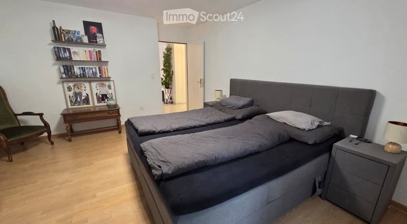 A well-lit bedroom with a large double bed, nightstand, wooden chair, small table with shelves, and decorative items.