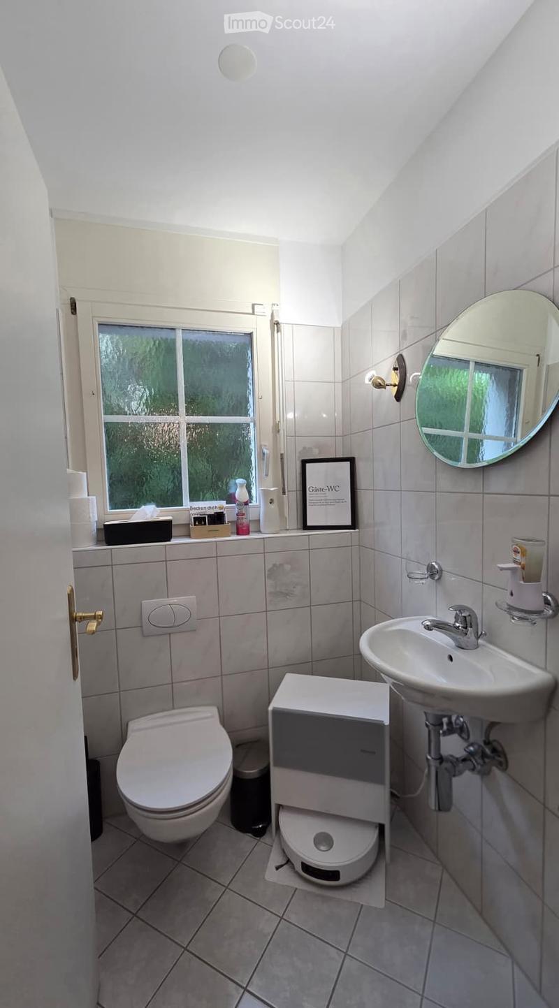 A small, modern bathroom with a toilet, sink, mirror, and a small cabinet. The room features tiled walls and floor, a window with a view of greenery, and various toiletries and cleaning supplies on the windowsill.