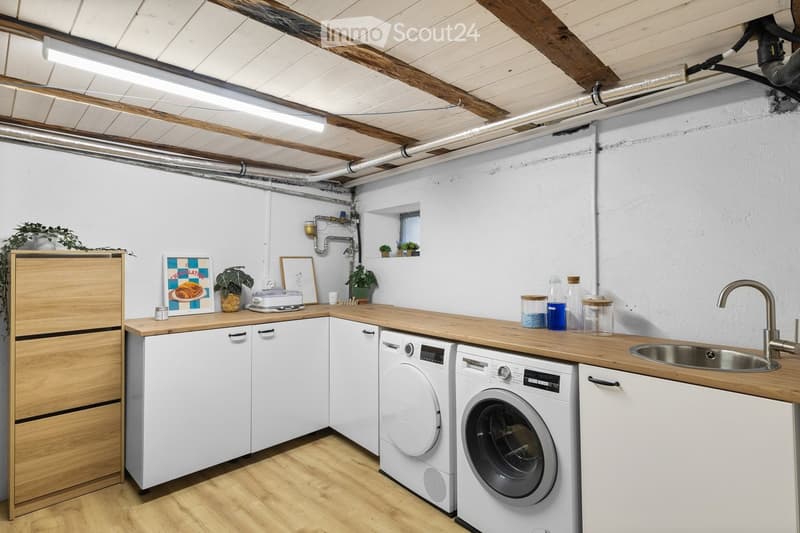 Modern kitchen with white cabinets, wooden countertops, stainless steel sink, and integrated washing machine and dishwasher. Features exposed wooden beams on the ceiling and small potted plants on the windowsill.