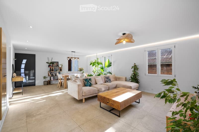 Modern living room with a large sectional sofa, wooden coffee table, dining area with a table and chairs, bookshelf, and several potted plants. The space features large windows and a sleek, contemporary design.
