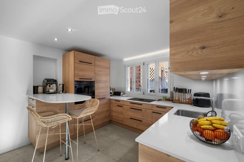 Modern kitchen with white countertops, wooden cabinets, and stainless steel appliances. Features a round island with a table and two wicker chairs, a bowl of fruit on the counter, and various appliances including a coffee maker, oven, and toaster.