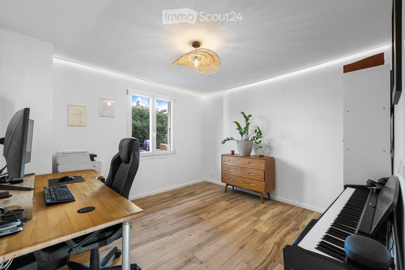 Modern office space with wooden flooring, large window, desk with computer setup, ergonomic chair, wooden cabinet, potted plant, and a small piano.