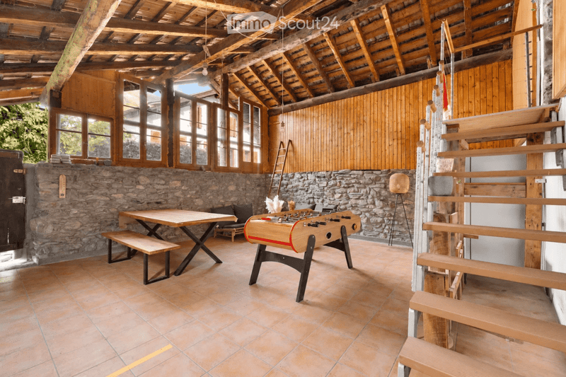 A rustic, open-plan living space with exposed wooden beams, stone walls, large windows, a wooden table with benches, a sofa, a foosball table, and a modern stool lamp.