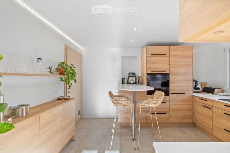 Modern kitchen with light wooden cabinets, white countertops, and a small round table with two wicker stools. The kitchen features a built-in oven, coffee maker, and various decorative items on shelves and countertops.