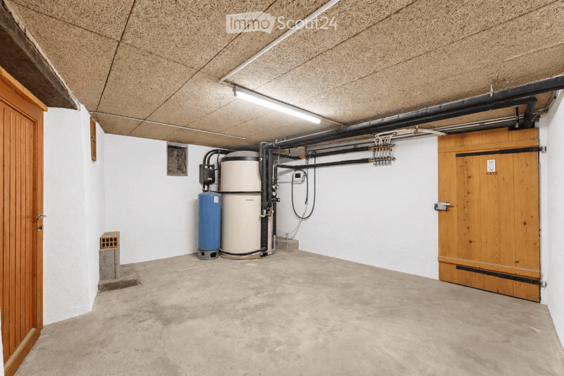 Empty utility room with concrete floor, white walls, exposed pipes, and a water tank. Wooden doors on both sides.