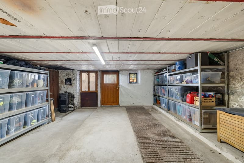 A spacious, unfinished basement or storage area with concrete walls and ceiling, featuring metal shelving units filled with storage bins and miscellaneous items, a wooden door, a radiator, and a small table.