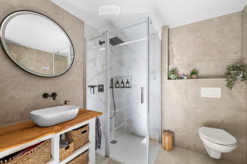 Modern bathroom with a white sink, wooden vanity, large round mirror, glass-enclosed shower, and a toilet. Features include a marble tile backsplash, wicker baskets under the vanity, and small decorative plants on the shelves.