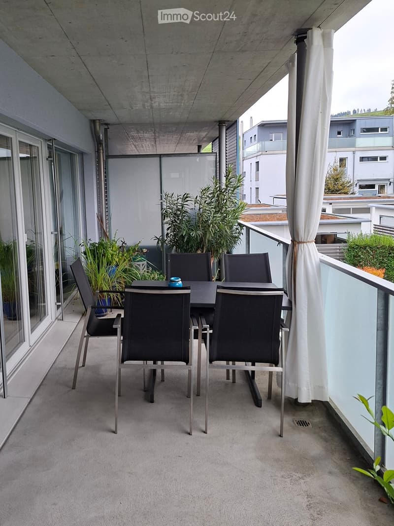 Modern balcony with a concrete floor, glass railing, and a dining set including a table and four chairs. Potted plants are placed around the area, and a white curtain is drawn on one side.