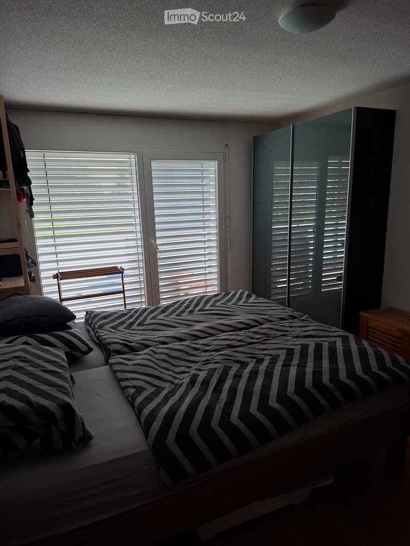 Bedroom with a double bed, pillows, a wardrobe, and a chair near the window with blinds.