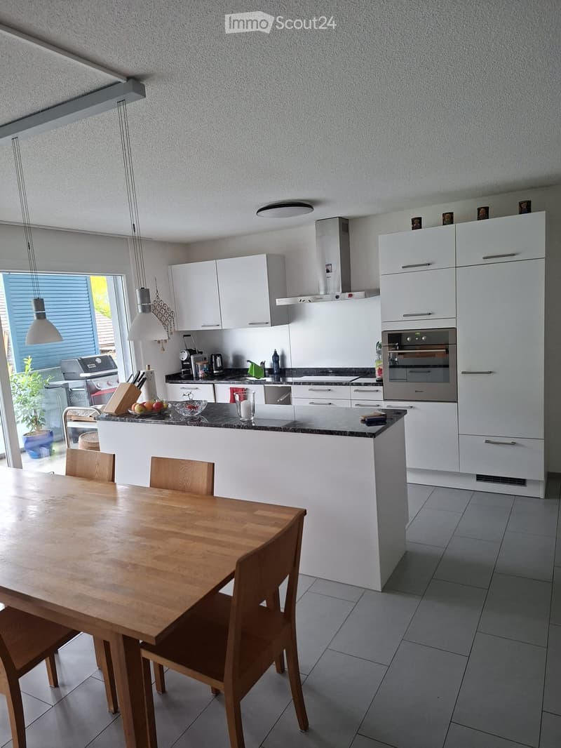 Modern kitchen with white cabinets, a central island, and a dining area. Features include a wooden dining table with chairs, a built-in oven, refrigerator, and various kitchen appliances.