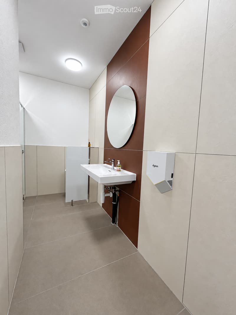 Modern bathroom with a white sink, round mirror, and sleek fixtures. Beige tiles and warm lighting.