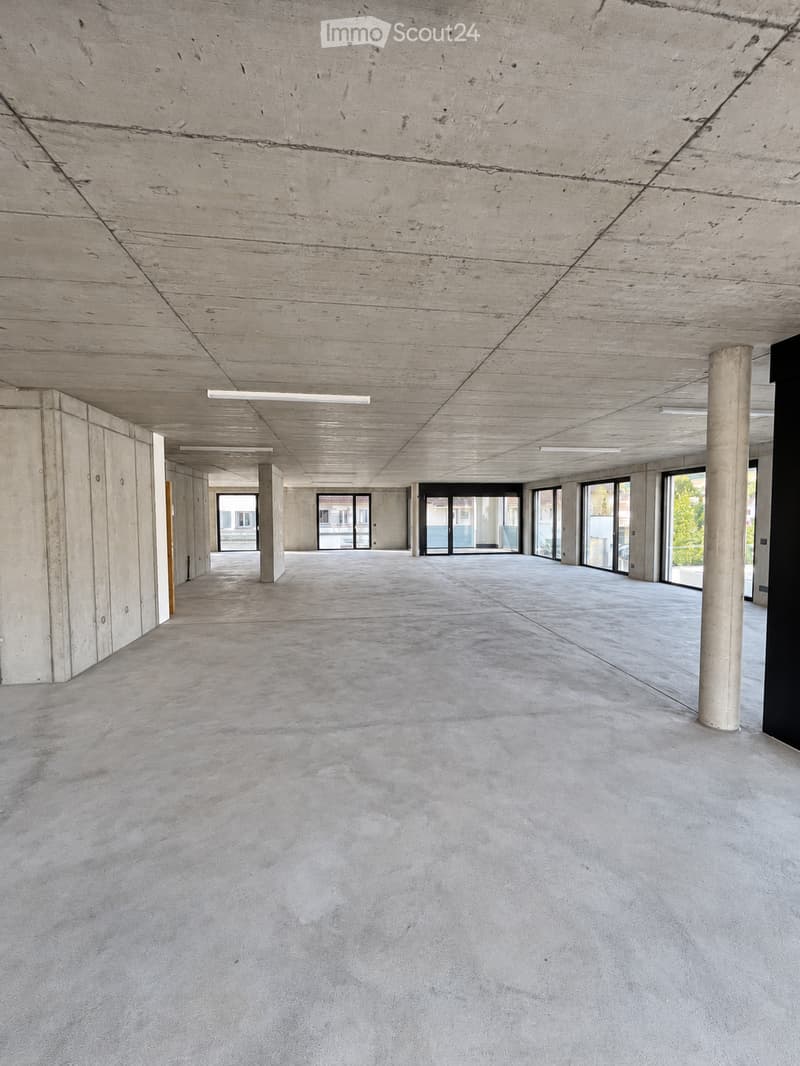Empty, modern corridor with concrete floors and walls, large windows at the end, and exposed ceiling beams.